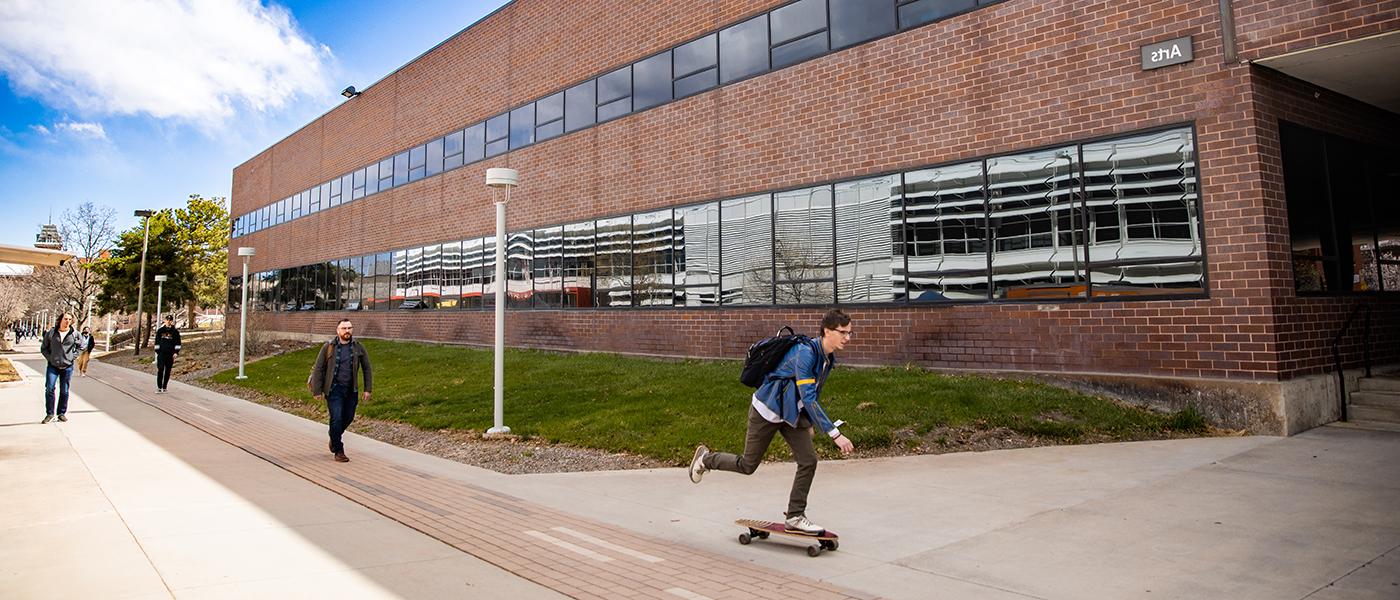 Brick building is the exterior of the Arts Building, and a student is skateboarding by.